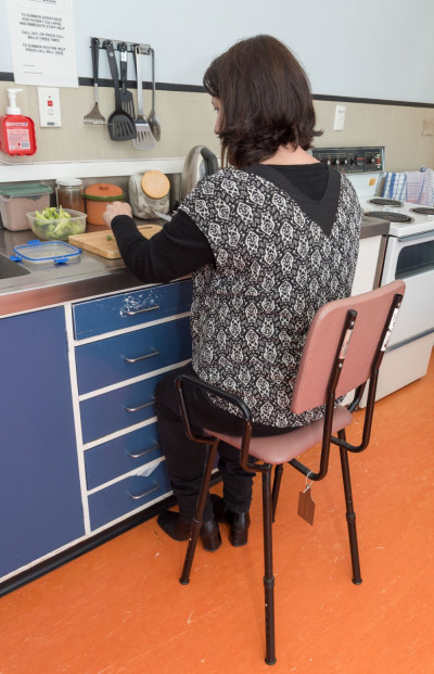 A woman sits on a tall stool with back and armrests while chopping vegetables at a kitchen bench. 