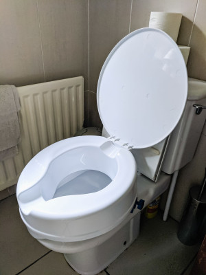A white plastic toilet seat about 10 centimetres tall that has been fixed over an existing toilet. the effect it to raise the height of the existing seat. 